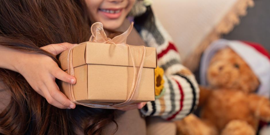 A child in a striped sweater handing a ribbon-tied gift box to their mom, a teddy bear in a Santa hat on the couch beside them